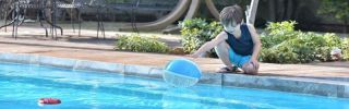 young boy leaning over edge of inground pool trying to reach a beach ball floating in the water.